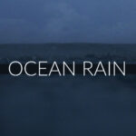 Rain falling over the open ocean with rippling water and moody skies.