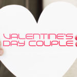 A man in a green hoodie and a woman in a red and cream sweater standing side-by-side, each holding a white paper heart in front of a white brick wall.