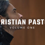 A man in a black suit walking solemnly through a traditional church sanctuary with wood-paneled walls and classic pews.